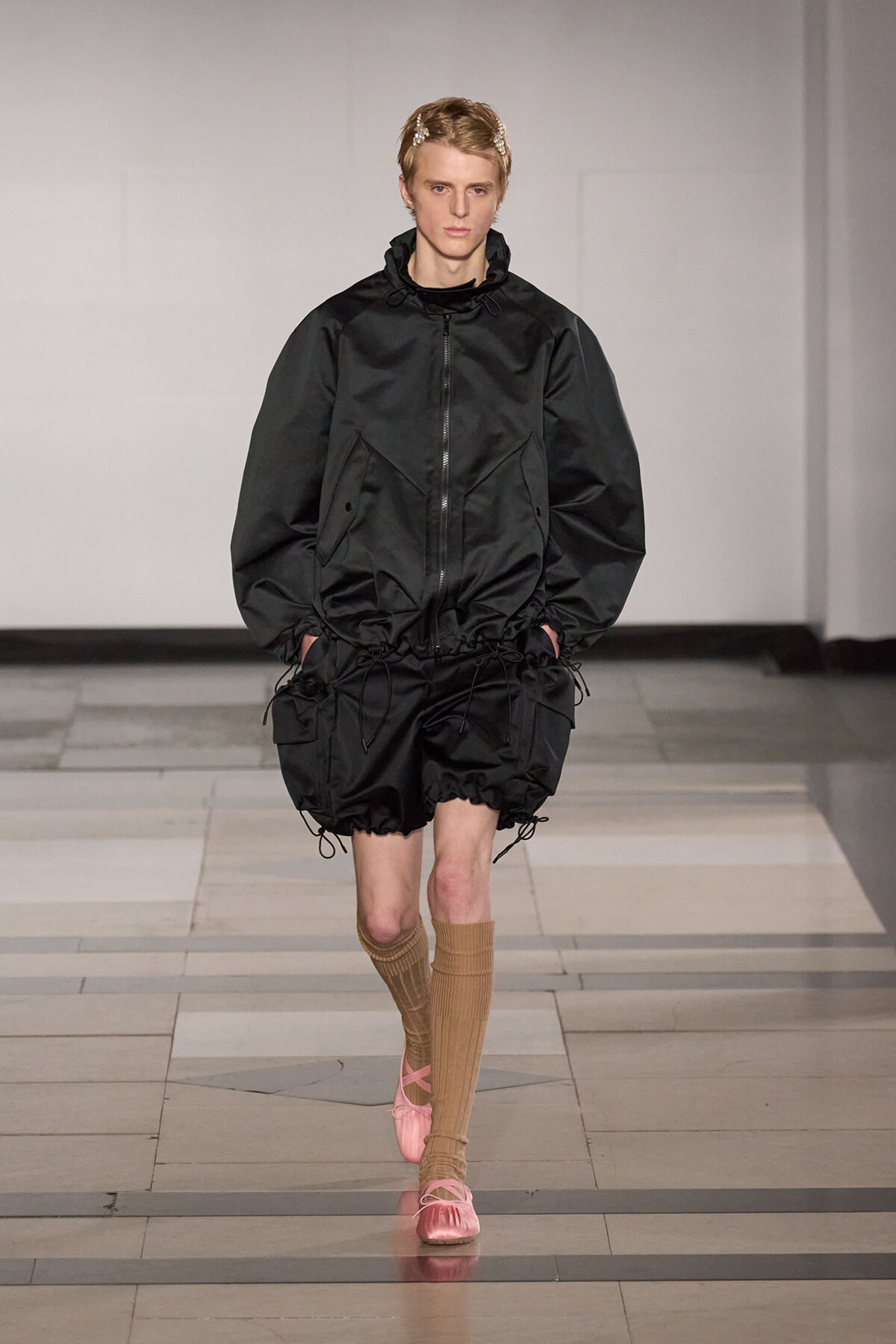 Model walks a fashion show in an oversized black windbreaker with drawstring hem, paired with black bubble shorts, tan knee-high socks, and pink ballet flats on a tiled runway.