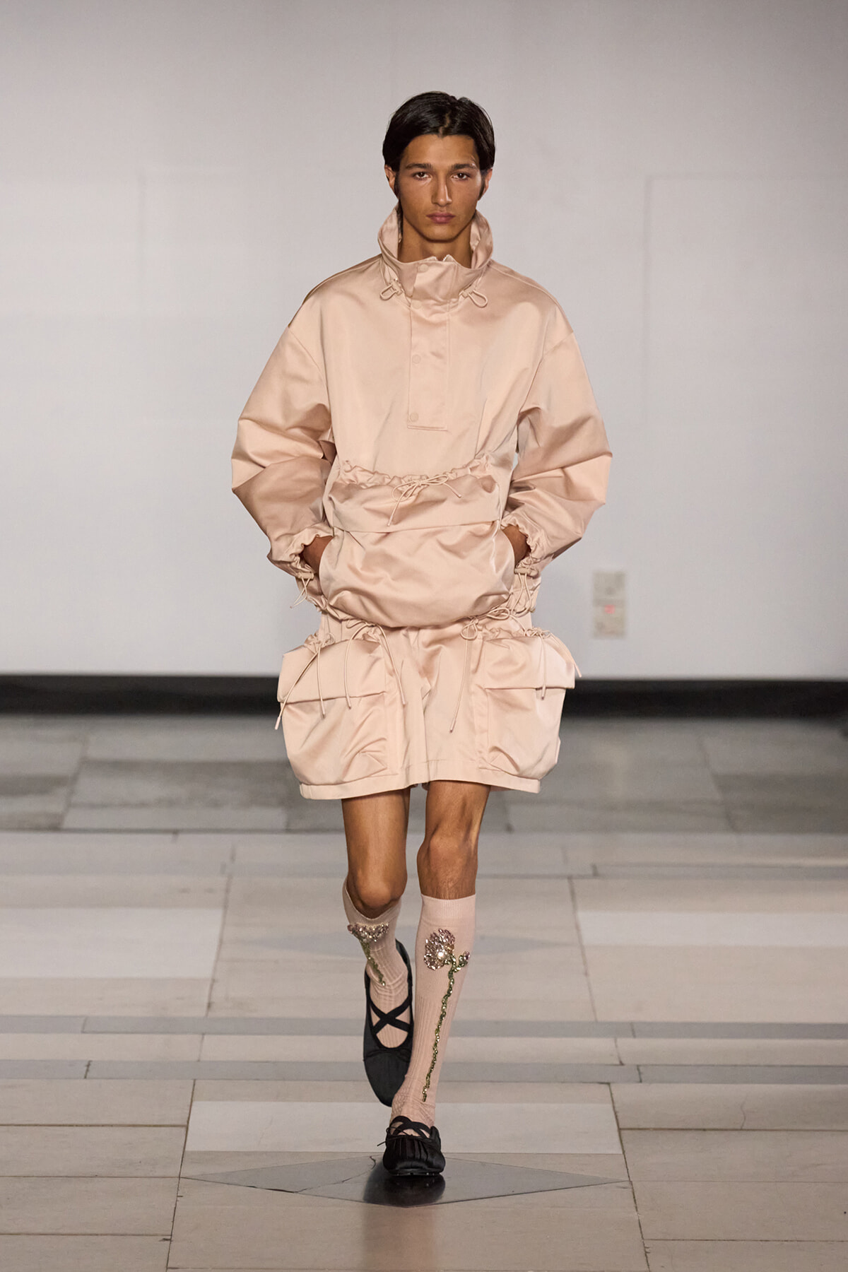 Model walking runway in a pale pink oversized windbreaker with drawstrings and matching shorts with large pockets and gathered hems due to a fashion-forward ensemble.