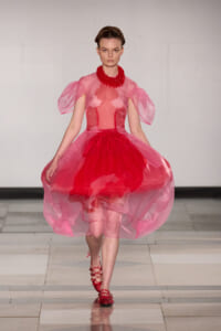 Model on a runway wearing a pink sheer-ballgown with a red layered skirt, puff sleeves, and a bold red statement necklace, red sandals visible down the catwalk.