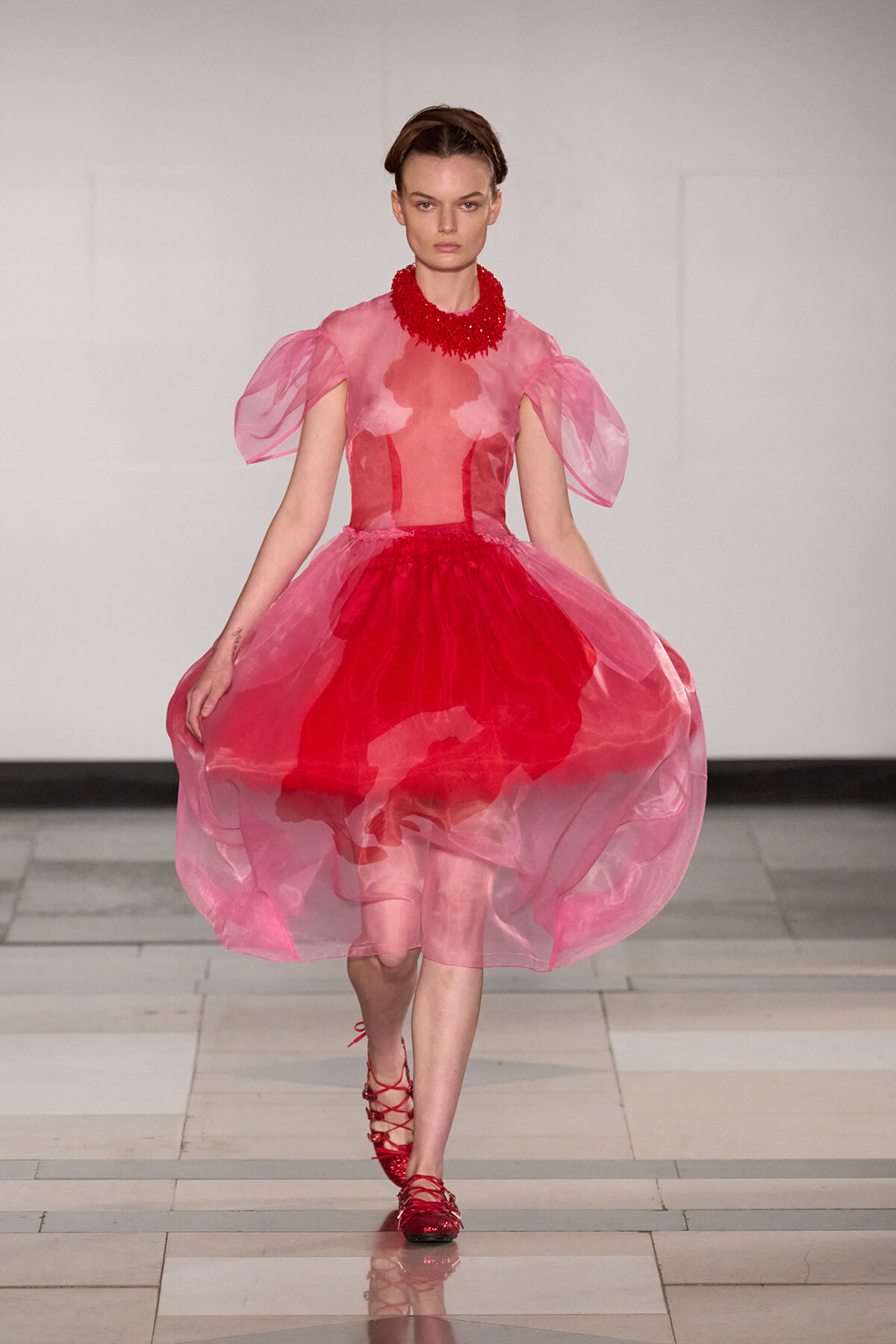 Model on a runway wearing a pink sheer-ballgown with a red layered skirt, puff sleeves, and a bold red statement necklace, red sandals visible down the catwalk.