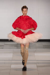 Model walking a runway in a bright red oversized top with a pink tutu skirt and embellished knee-high socks; sleek hair, poised pose.