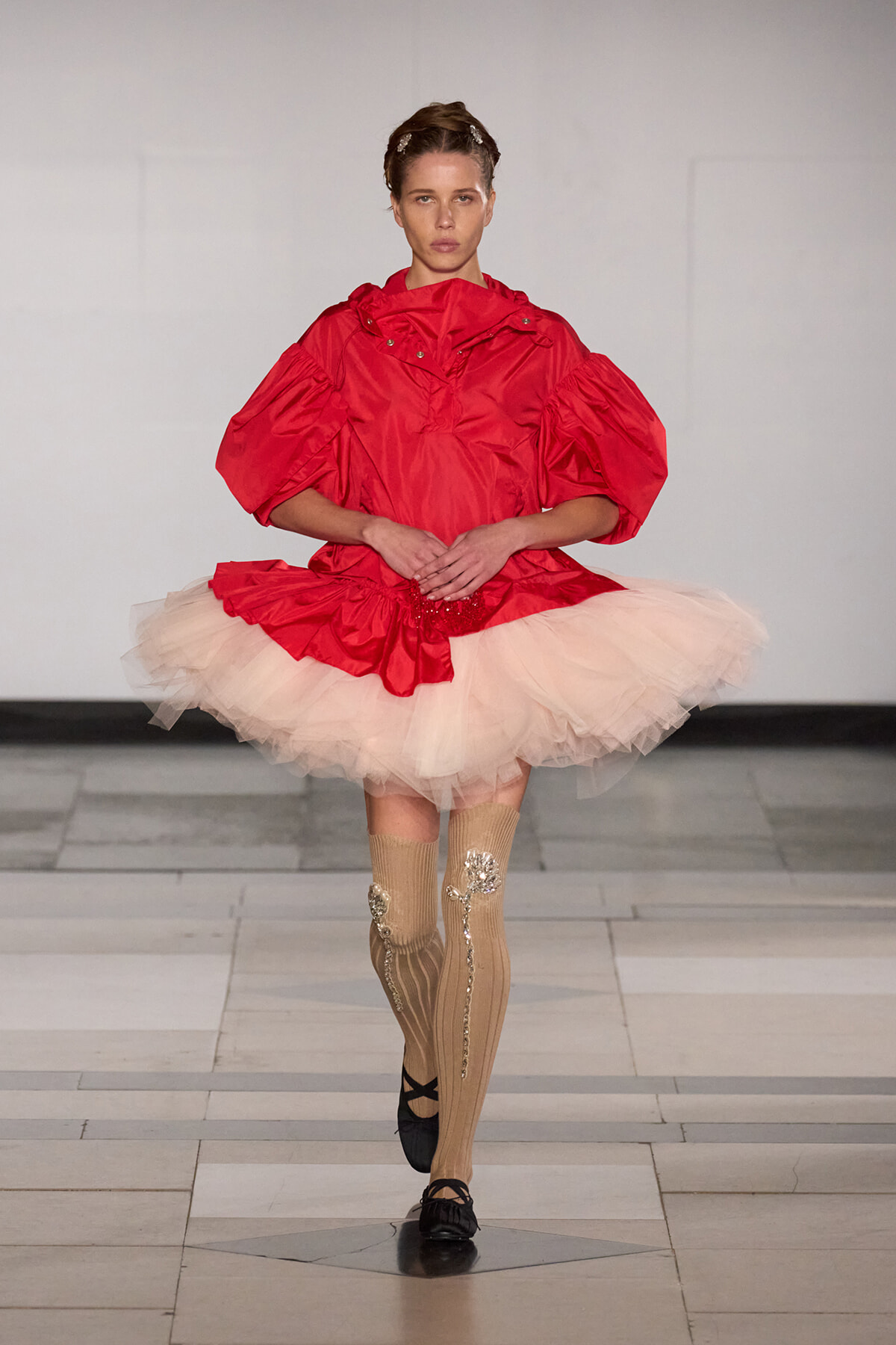 Model walking a runway in a bright red oversized top with a pink tutu skirt and embellished knee-high socks; sleek hair, poised pose.
