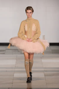 Model walks the runway in a beige cardigan over a light pink ballet tutu and matching undergarments; knee-high embellished socks and dark dress shoes complete the look.