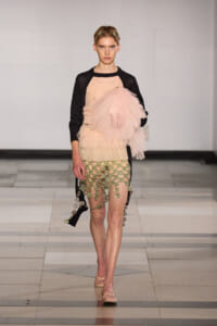 Model walking in a pink layered tulle skirt with a black cardigan on a runway, pink ballet flats visible.