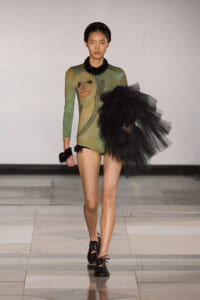 Model walks the runway in a green bodysuit with a face print, paired with a dramatic black tulle cluster on one side and chunky black jewelry.