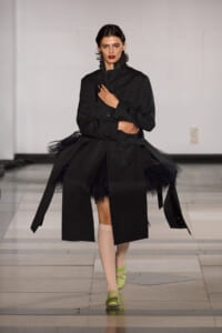 Model walking the runway in an oversized black coat with tulle underskirt, beige socks, and neon-green shoes; bold red lipstick and short dark hair.