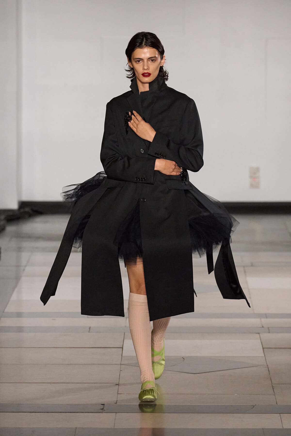 Model walking the runway in an oversized black coat with tulle underskirt, beige socks, and neon-green shoes; bold red lipstick and short dark hair.