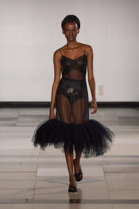 Model walking a runway in a sheer black lingerie dress with a voluminous tulle skirt and braided hair.