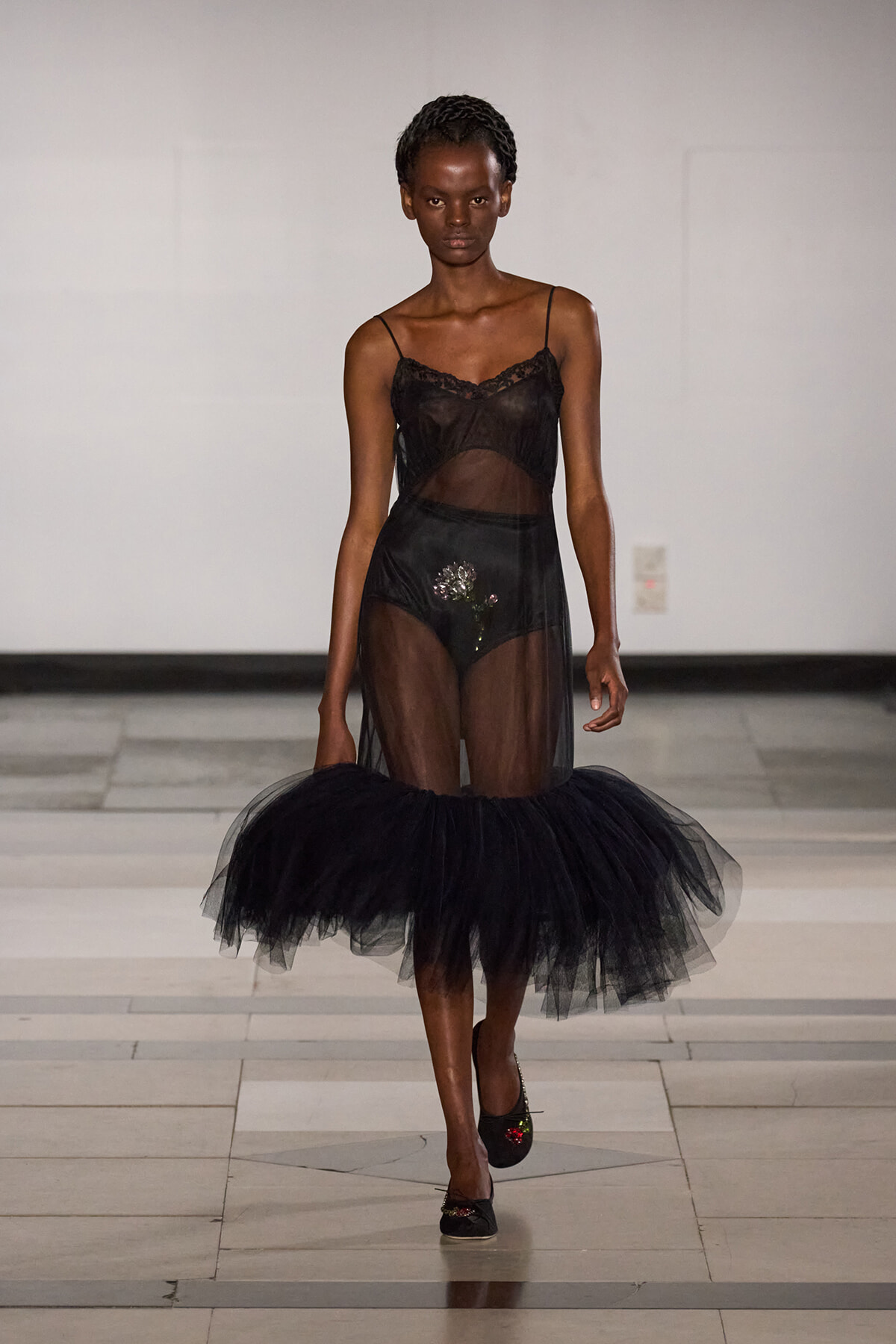 Model walking a runway in a sheer black lingerie dress with a voluminous tulle skirt and braided hair.