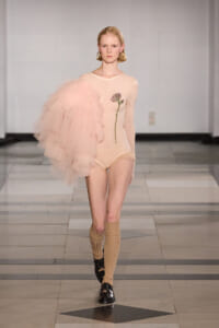 Model walks the runway in a pale pink sheer bodysuit with a large pink tulle sleeve on the left, plus a floral appliqué along the side; beige knee-high socks and black shoes.