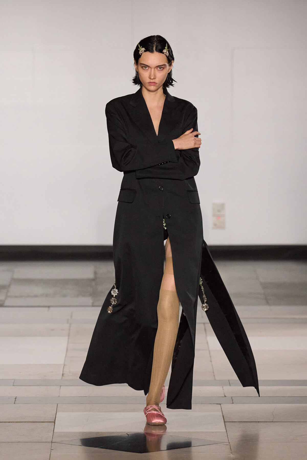 Model walks a runway in a long black coat with a front slit, revealing beige stockings and pink shoes; hair adorned with decorative clips.