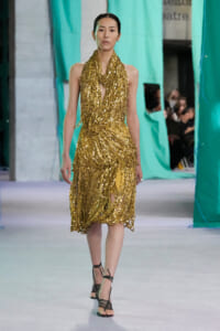 Model walking the runway in a gold sequined halter dress with layered drape details against teal backdrop.