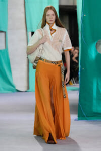 Model walking a runway in orange wide-leg pants and a white mesh top with orange trim, carrying a white bag over her shoulder at a fashion show.