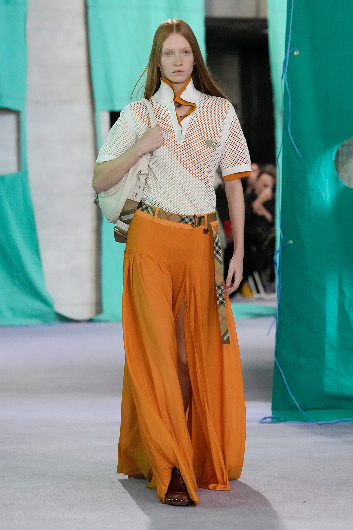 Model walking a runway in orange wide-leg pants and a white mesh top with orange trim, carrying a white bag over her shoulder at a fashion show.