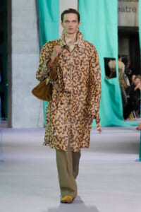 Model walking a runway in a long brown floral coat, beige pants, yellow shoes, and a brown shoulder bag.