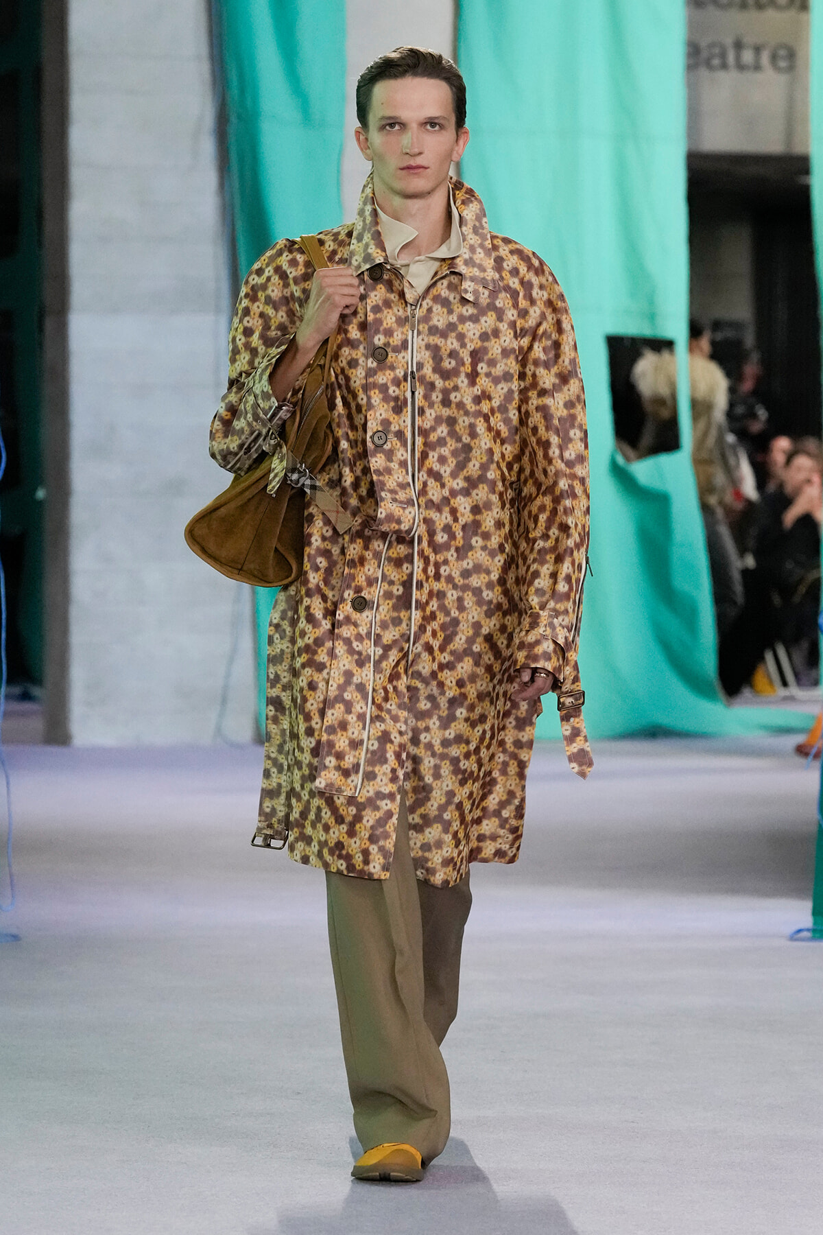 Model walking a runway in a long brown floral coat, beige pants, yellow shoes, and a brown shoulder bag.
