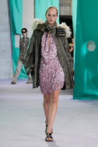 Model walking a runway in a pink sequin mini dress under an oversized olive-green parka with fur-trimmed shoulders.