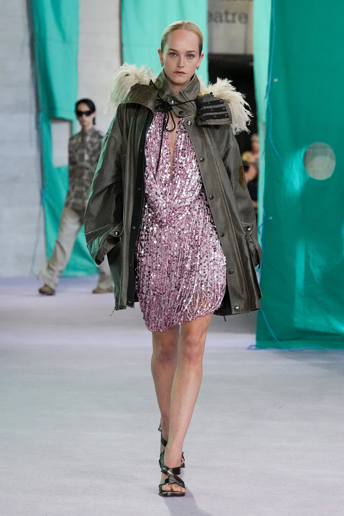 Model walking a runway in a pink sequin mini dress under an oversized olive-green parka with fur-trimmed shoulders.