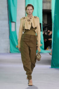 Female model on a fashion runway wearing a beige cropped trench with buckle straps over an olive top and high-waisted khaki cargo pants; carrying a small metallic bag and wearing strappy heels.