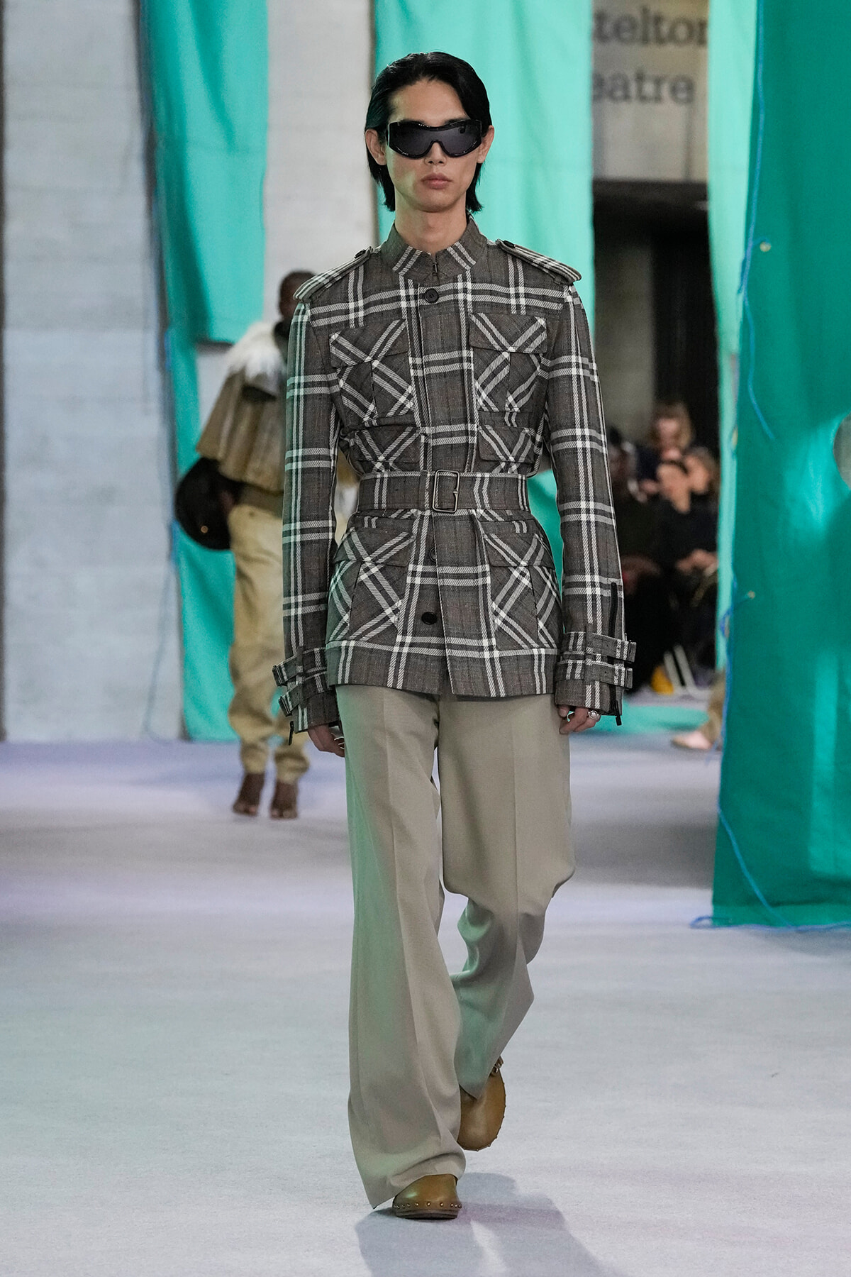 Model walking the runway in a gray plaid belted jacket with wide beige trousers and dark sunglasses.