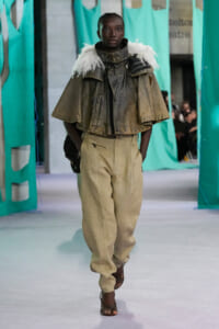 Model walks a fashion runway wearing a khaki cape with white fur trim over a rugged jacket and high-waisted beige trousers