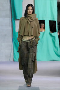 Model walking a fashion runway in an oversized olive cape with a high, sculpted collar and a draped scarf detail, paired with green-brown plaid baggy pants and a metal chain belt.