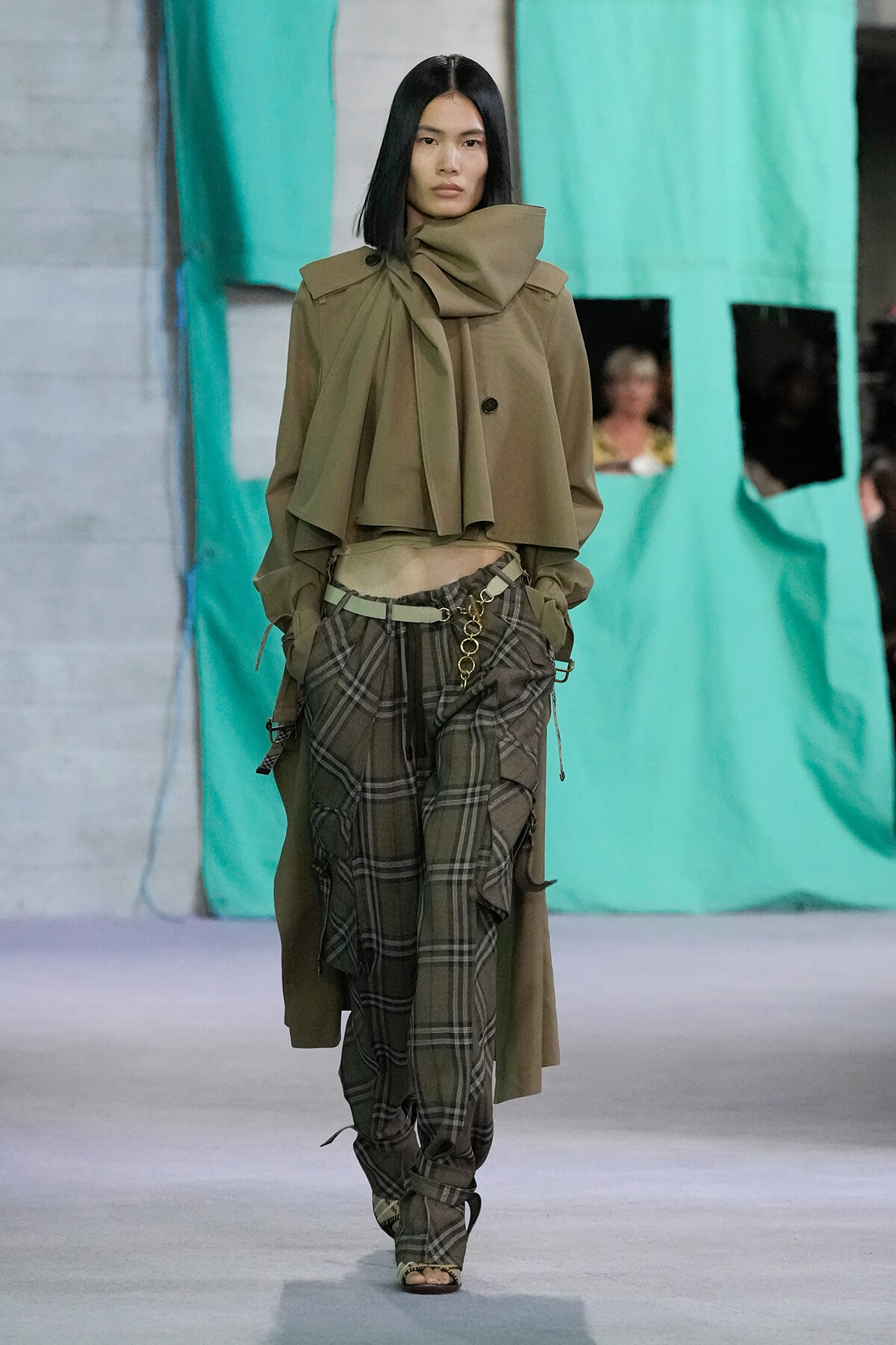 Model walking a fashion runway in an oversized olive cape with a high, sculpted collar and a draped scarf detail, paired with green-brown plaid baggy pants and a metal chain belt.