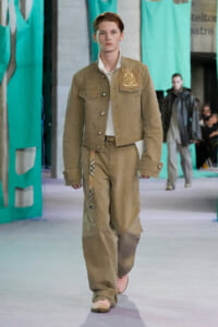 Male model walks a fashion runway wearing a beige utility jacket with a crest patch and matching wide pants; pink sneakers visible, teal fabric backdrops behind.