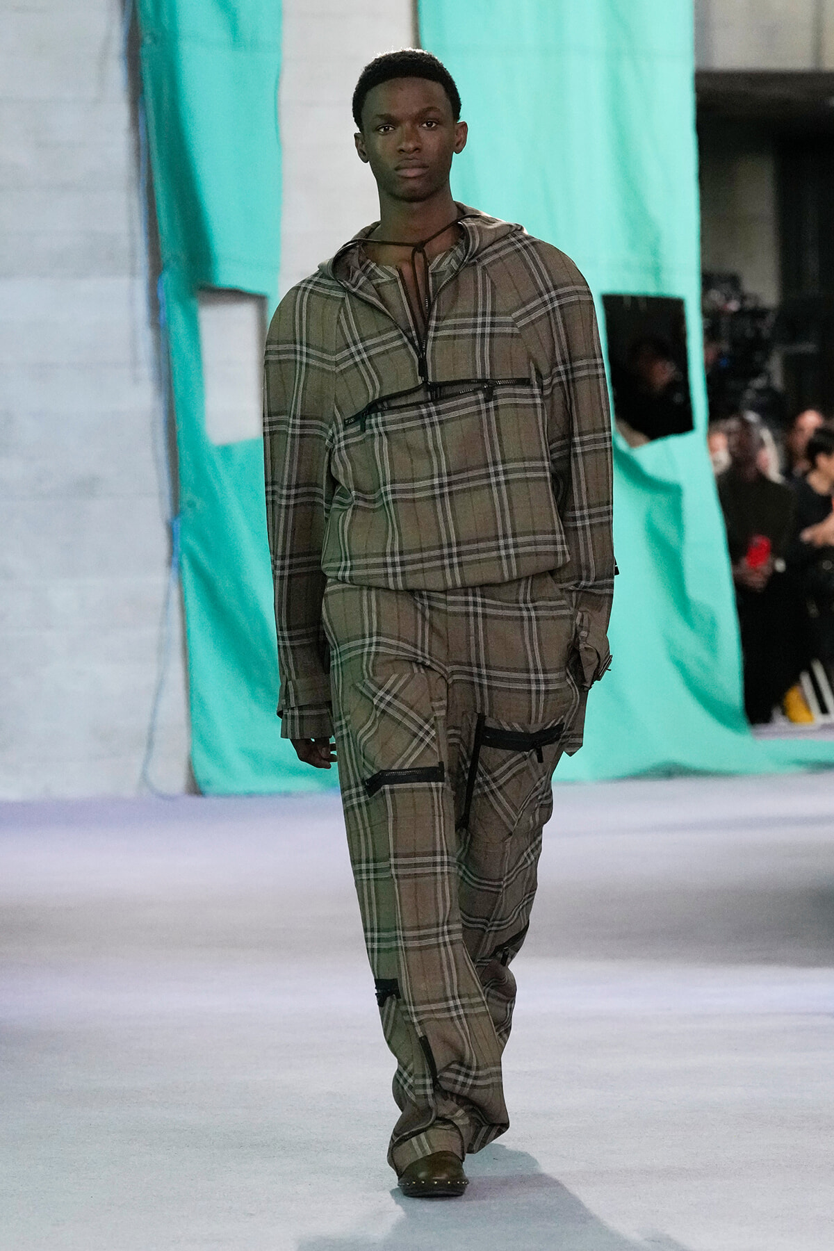 Male model walking runway in a brown plaid zip-up top and matching pants with large pockets, teal backdrop behind him.