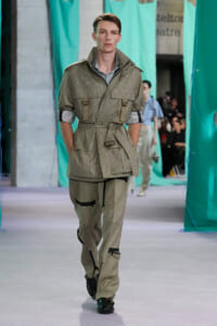 Male model walks the runway in a beige utilitarian jacket with multiple pockets and matching pants, belt cinching the waist.