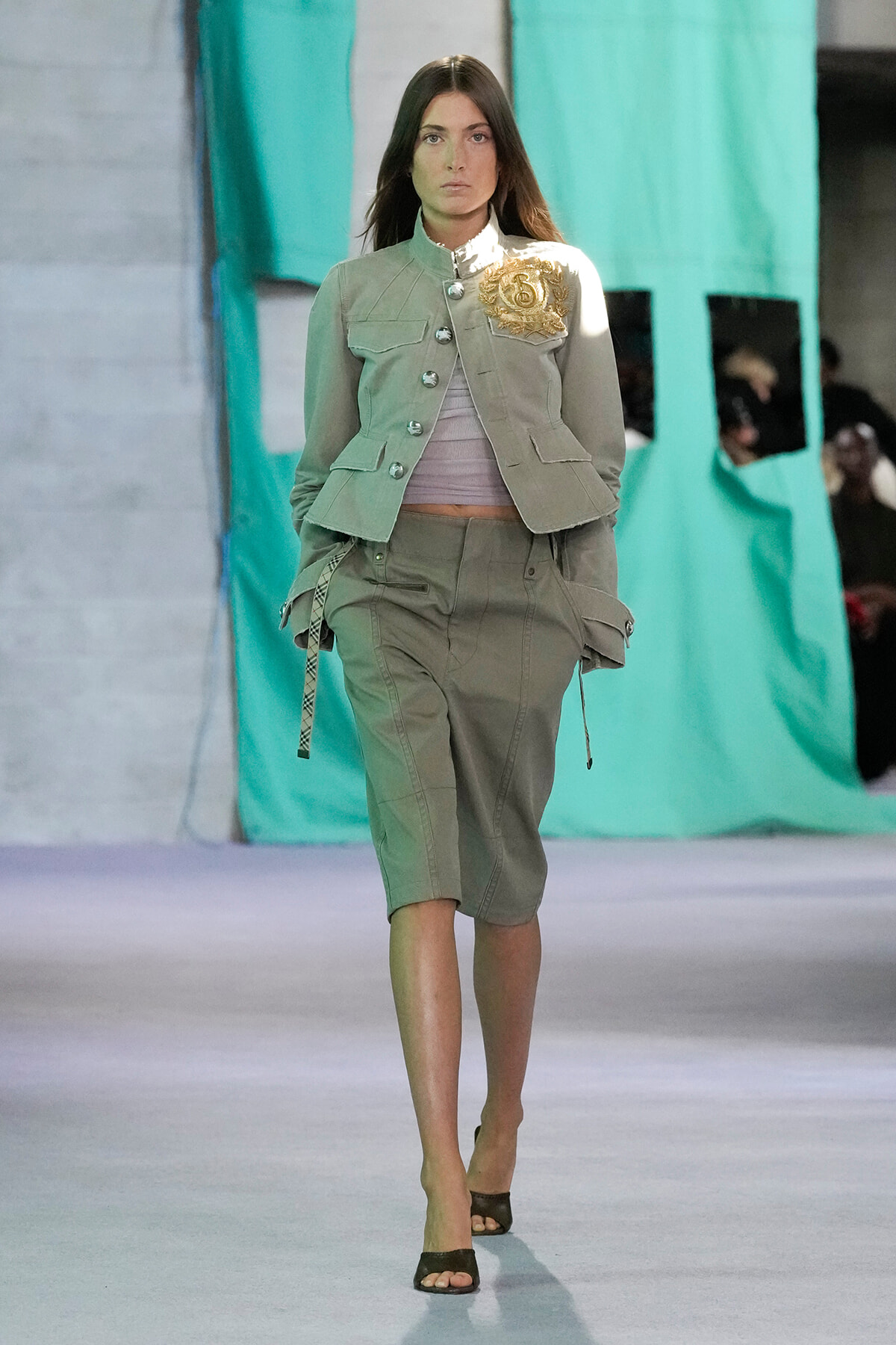 Female model walking a runway in a beige utility jacket with gold crest patch and matching cargo shorts, open-toe heels.