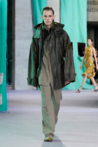 Male model walking a runway in an oversized dark green leather jacket with epaulets over a beige suit.