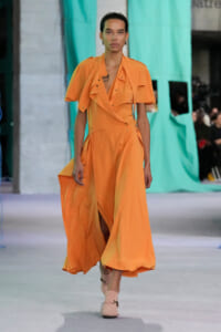 Model walking a runway in a bright orange draped jumpsuit with wide legs and short sleeves, paired with pale pink sandals.