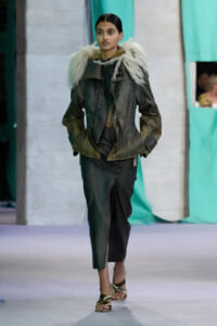 Model walking runway in a dark green jacket with a white faux fur collar, matching green pants, and strappy sandals.