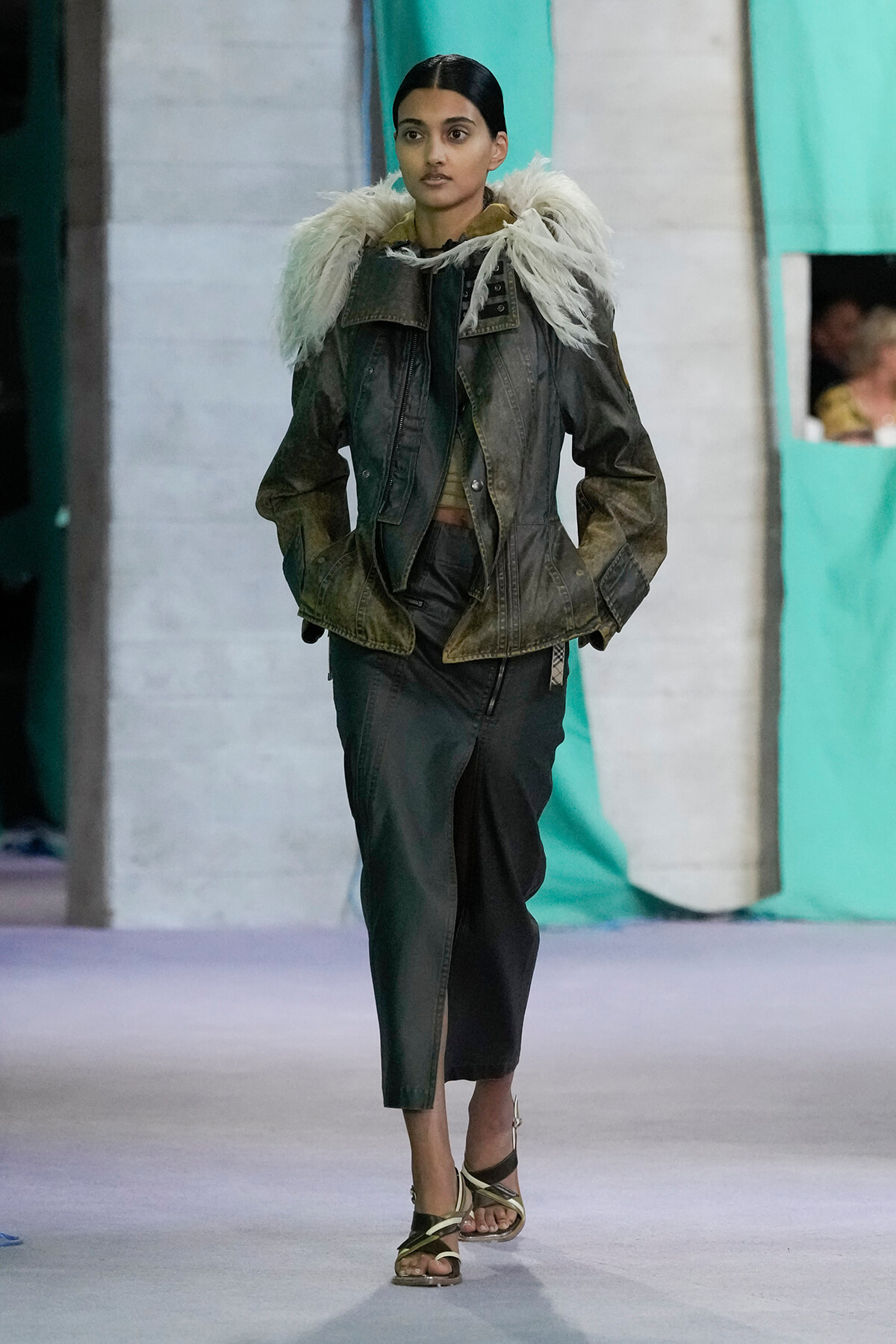 Model walking runway in a dark green jacket with a white faux fur collar, matching green pants, and strappy sandals.