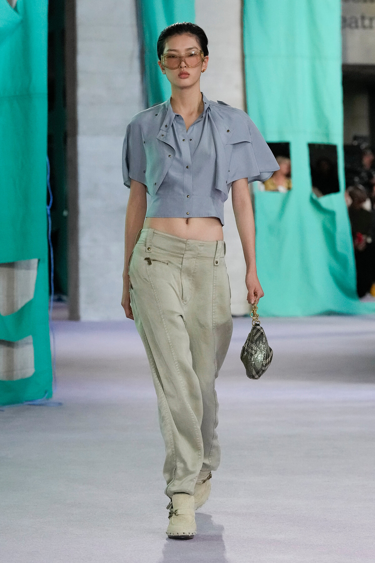 Model on a runway wearing a light blue cropped jacket, beige pants, beige boots, tinted sunglasses, and a small metallic handbag.