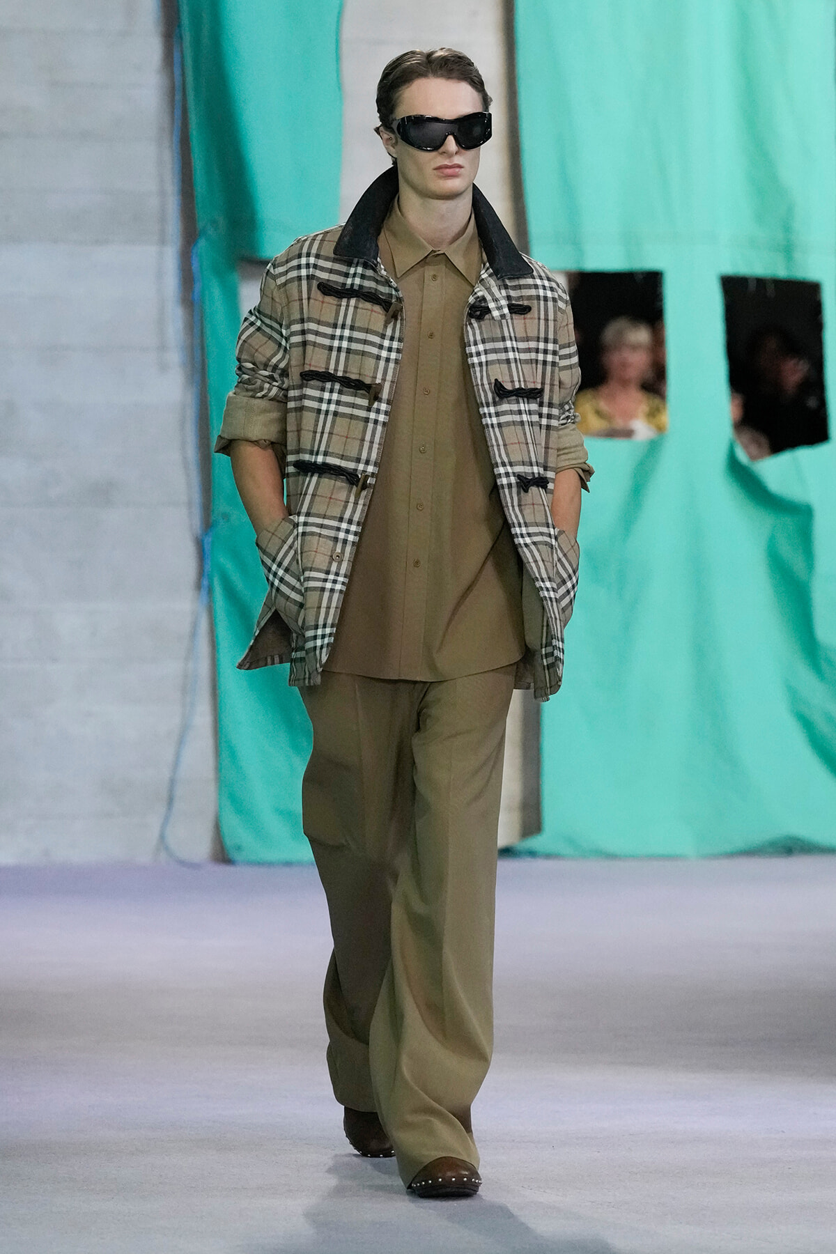 Model on a runway wearing a beige shirt and trousers under a plaid jacket with rolled sleeves, dark sunglasses.