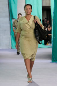 Model walking a fashion show in a beige dress, carrying a large black handbag over her shoulder.