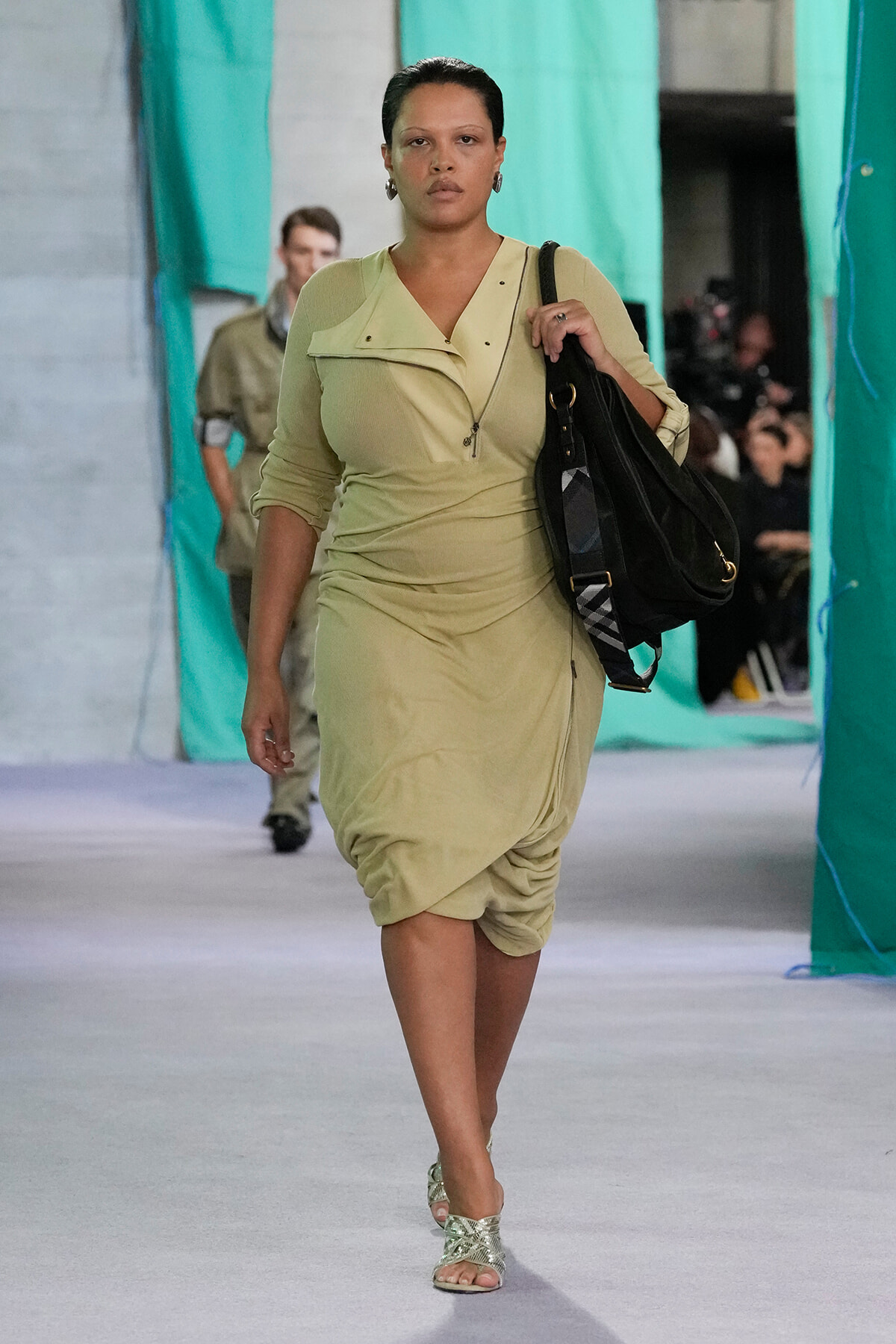 Model walking a fashion show in a beige dress, carrying a large black handbag over her shoulder.