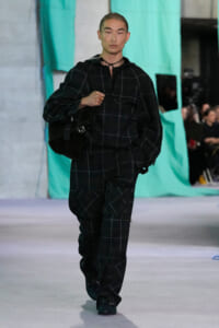 Male model walking a runway in a black plaid oversized outfit, carrying a large black bag against a teal backdrop.