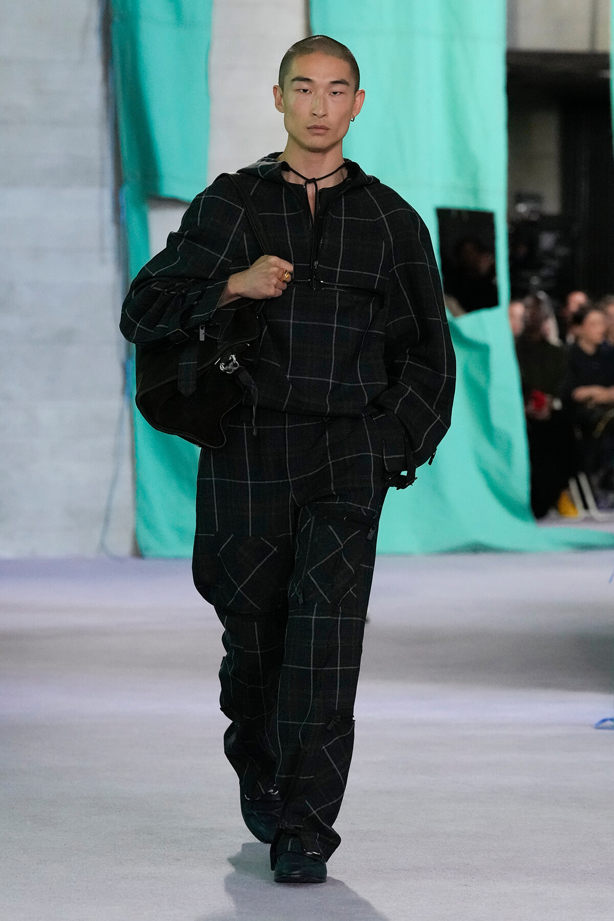Male model walking a runway in a black plaid oversized outfit, carrying a large black bag against a teal backdrop.