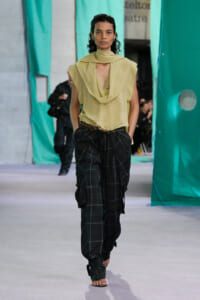 Model walks a runway in a pale yellow draped sleeveless top with a scarf-like collar and dark plaid baggy pants with a belt.