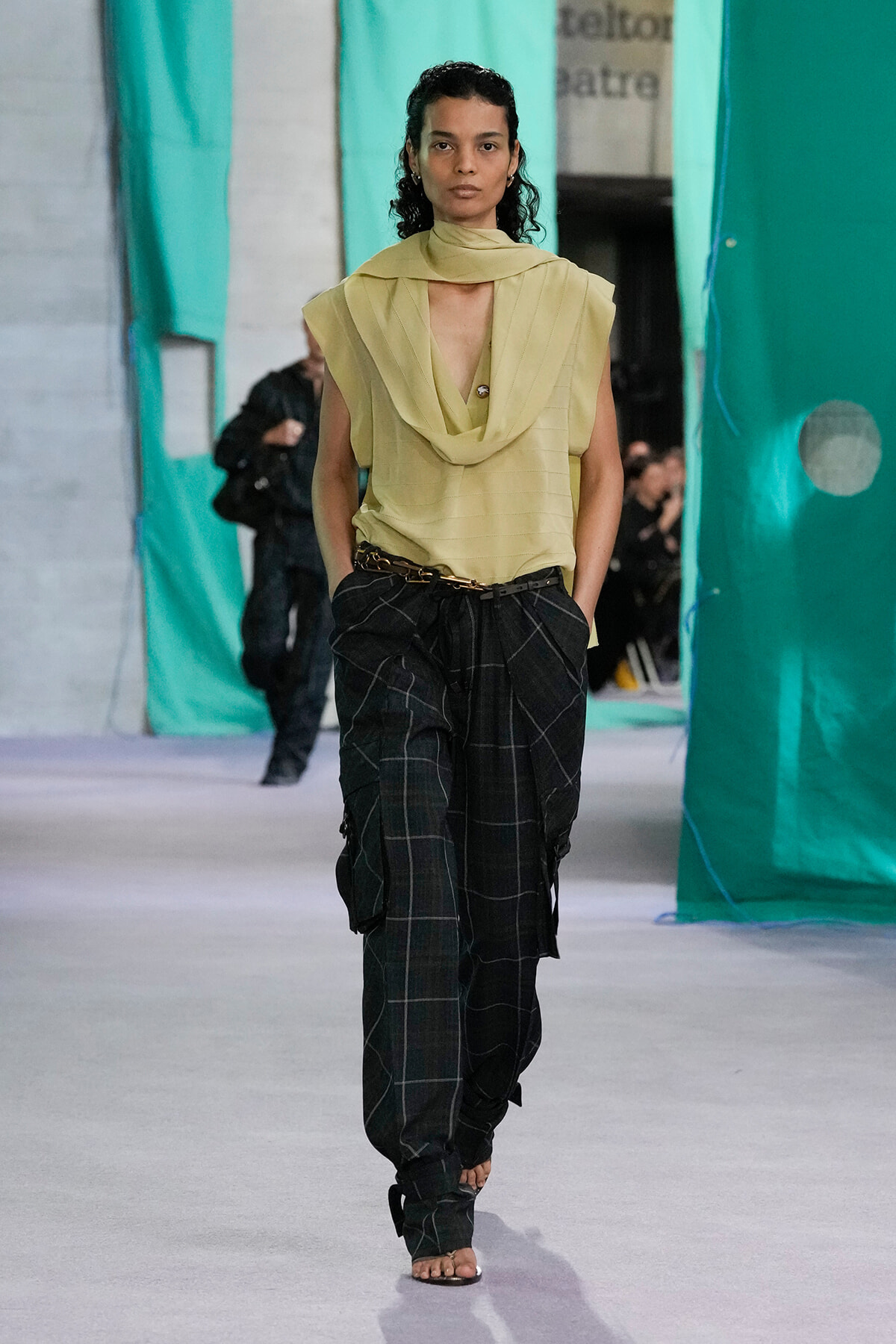 Model walks a runway in a pale yellow draped sleeveless top with a scarf-like collar and dark plaid baggy pants with a belt.