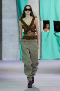 Model on a runway wearing a brown sequin ruffled top, olive cargo pants, and purple ankle boots, with oversized sunglasses.