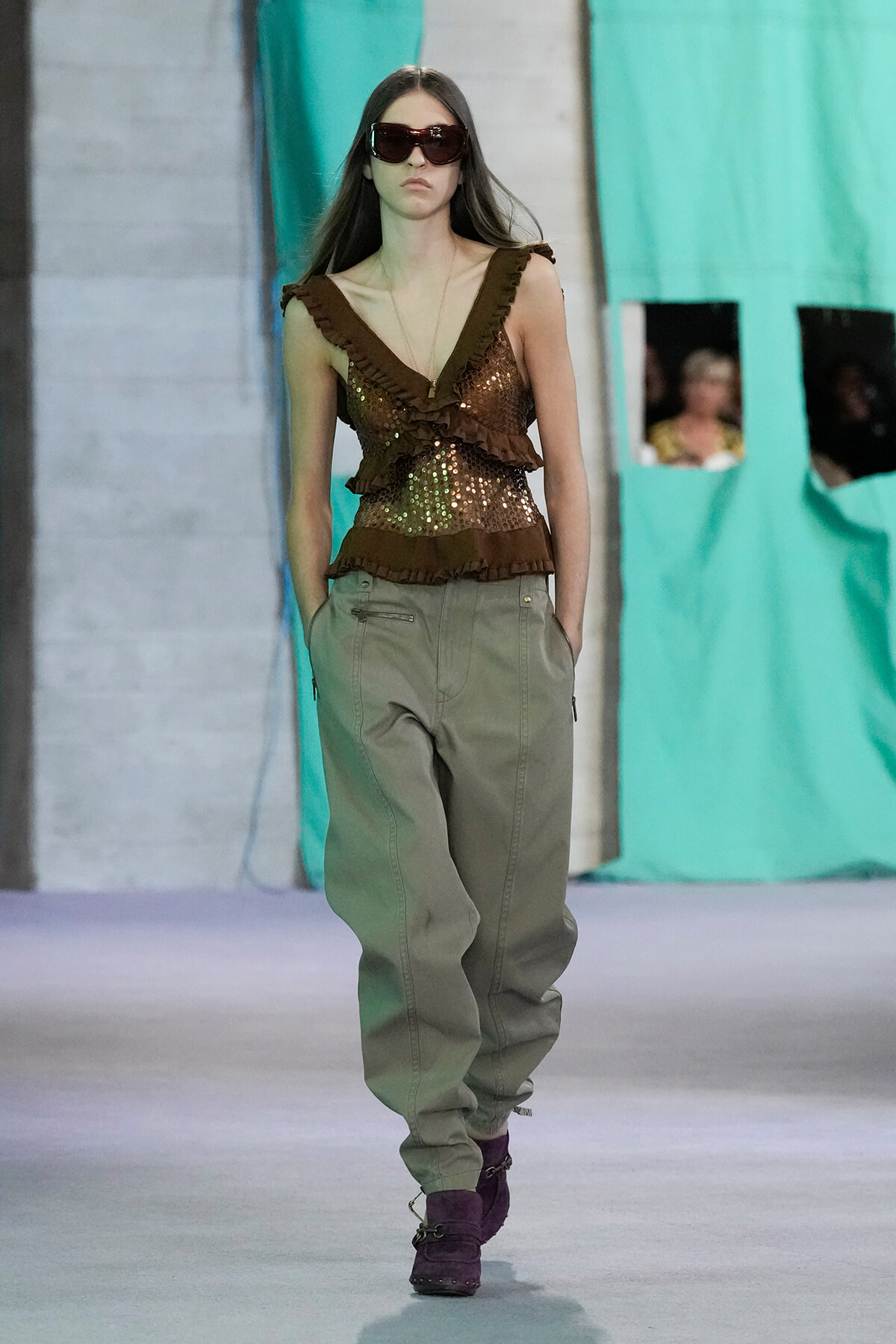 Model on a runway wearing a brown sequin ruffled top, olive cargo pants, and purple ankle boots, with oversized sunglasses.