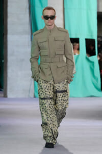 Male model on a runway wearing a beige military-style jacket with multiple pockets and camouflage leopard-print cargo pants, sunglasses on.