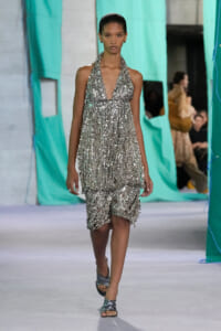 Model walking a runway in a silver sequined halter dress with layered fringe and metallic sandals, teal backdrop behind.
