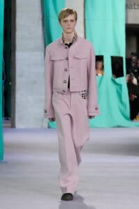 Model on a runway wearing a pale pink tailored jacket with large pockets and matching wide-leg pants, plaid shirt at the neck showing.
