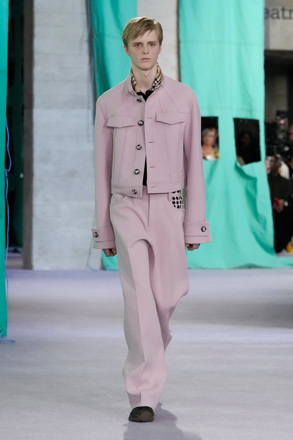 Model on a runway wearing a pale pink tailored jacket with large pockets and matching wide-leg pants, plaid shirt at the neck showing.