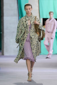 Female runway model walking in a pale floral trench over a lavender ruffled dress, carrying a brown satchel and wearing strappy sandals.